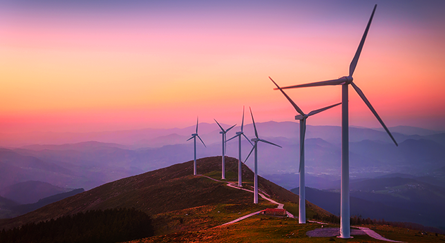 A photo of a row of wind turbines on a mountaintop against a backdrop of hazy purple mountains and a yellow and pink sunset