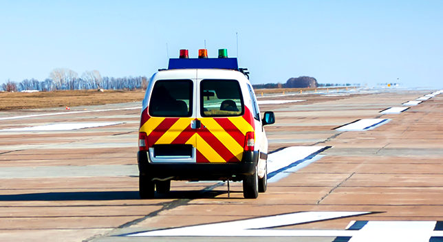 A photo of a utility van with red and yellow safety stripes painted on the back, driving along an airport runway
