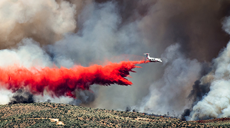An airplane flying through dark clouds of smoke dropping red flame retardant on a hillside