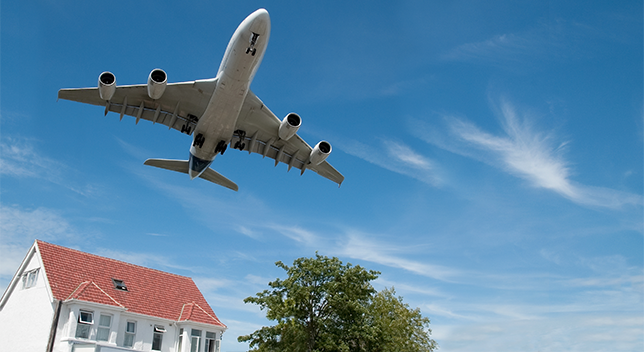 A photo of an airplane in a bright blue sky, flying low over a tree and a white house with a red roof
