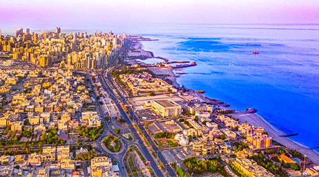 An aerial photo of a seaside city with warm yellow-toned buildings beside a vivid blue ocean against a pale blue and pink sky