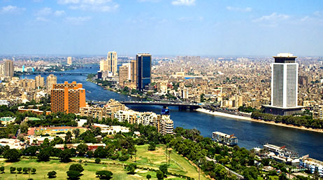 A modern city with white and beige buildings bisected by a blue river under a cloud-dotted blue sky