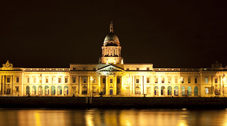 A stately government building brightly lit in gold tones against a night sky, mirrored in the still water of a nearby river
