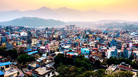 A colorful city with buildings in blue, red, and white in a tree-filled landscape against a backdrop of hazy blue mountains under a yellow and orange sunrise sky