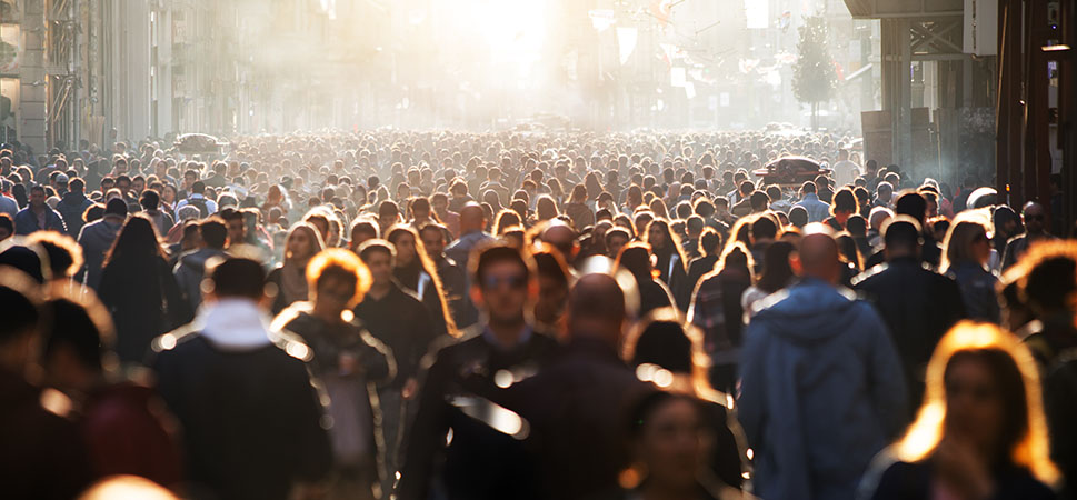 A photo taken over the heads of a large crowd of people on a city street, all silhouetted as they look in the direction of a bright light in the distance