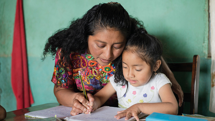 A woman hugging a child and guiding her hand as she practices writing letters in a softly lit classroom