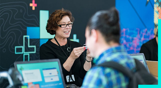 A photo of a conference staffer helping a registrant, while a person in a backpack stands in the foreground using a laptop