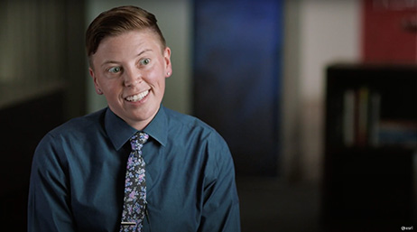 A screencap from the featured video with a smiling person in a blue collared shirt and a patterned purple tie in mid-conversation against a blurry dark background