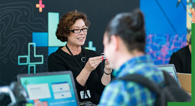 A photo of a conference staffer helping a registrant, while a person in a backpack stands in the foreground using a laptop