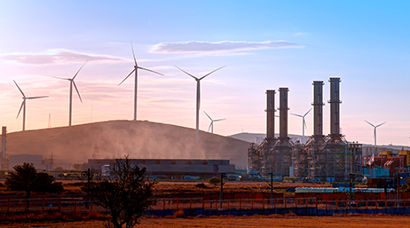An industrial complex beside several hills lined with wind turbines silhouetted against a clear blue sky