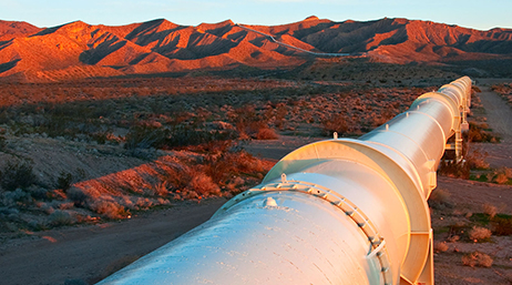 A large white pipe leads into the distance in a desert landscape with mountains lit rich orange by an unseen sunset