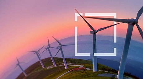 A green hillside with a row of white wind turbines against a purple mountainous horizon under a pink and orange sunset sky