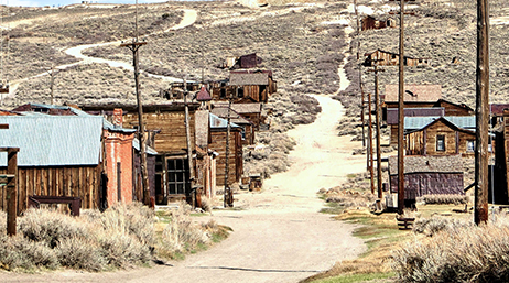 A dirt road trails through a historic mining town with weathered log buildings in a desert landscape of dry brown brush