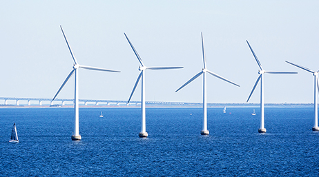 A row of white wind turbines in clear blue ocean waters with a white bridge in the distant background beneath a pale blue sky