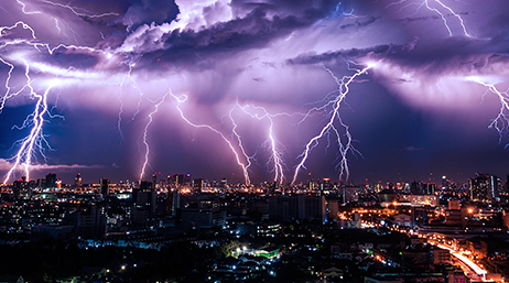 A brightly lit city at night under a stormy blue and purple sky filled with lightning bolts in mid-strike