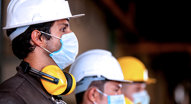 Three people in hard hats and face masks with noise-cancelling headphones around their necks standing in a row facing an unseen speaker on the right