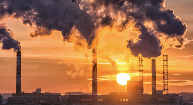 A factory with smoke billowing from several tall vertical pipes silhouetted against an orange and pink sunset