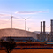 Photo of a brown field of cropland scattered with trees and topped with several wind turbines and factory smokestacks silhouetted against a pale afternoon sky