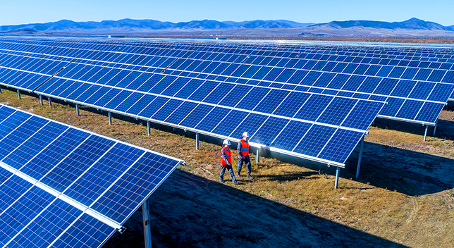 Photo of a field full of long stretches of blue solar panels with mountains in the distance as two people in orange safety vests and hard hats walk between the panels