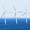 A row of wind turbines standing out of calm blue ocean waters beside a sailboat with a long white bridge in the distance