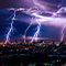 A city brightly lit for night under a stormy blue and purple sky with multiple lightning bolts in mid-strike between the clouds and the ground