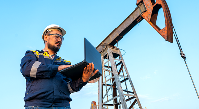 A photo of a utility worker in a hard hat holding an open laptop with a pumpjack in the background beneath a clear blue sky