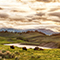 A rolling green prairie with grazing bison beside a shining river beneath a cloud-swept sunset sky