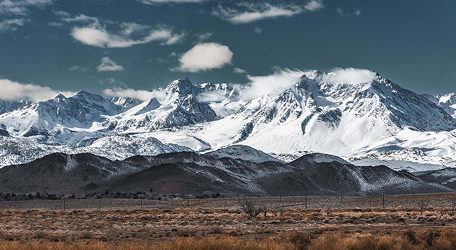 A snow-covered mountain range beneath a clear blue sky with a brown desert plain in the foreground