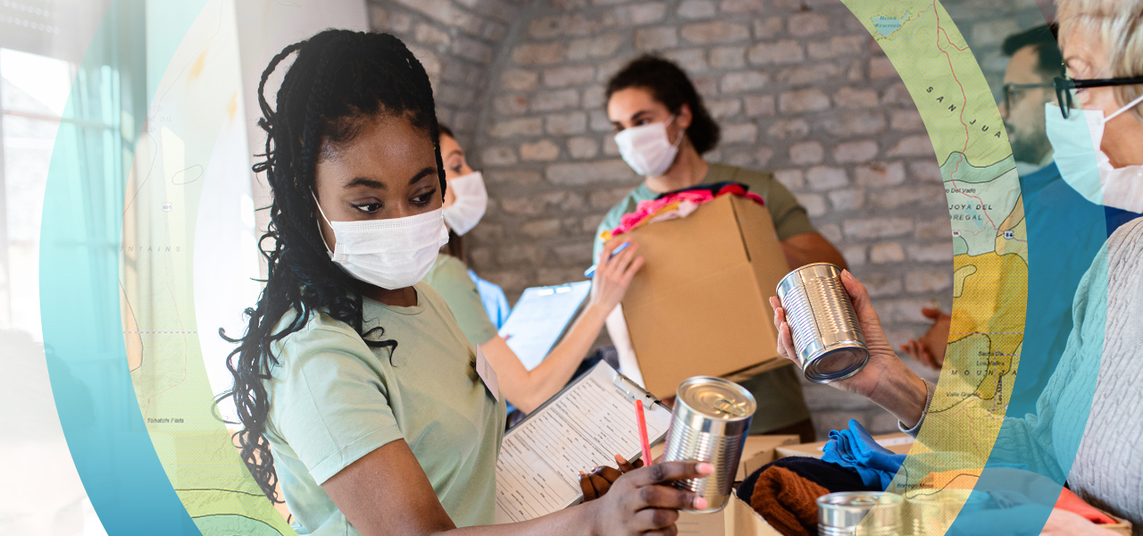 A photo of five relief workers in white face masks sorting boxes of clothing and canned food donations in a sunlit room with brick walls