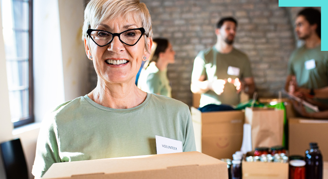 A photo of a smiling person in a pale green shirt and name badge holding a box in a brick-walled room, with several other volunteers standing near stacks of boxes in the background