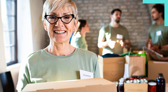 Una foto de una persona sonriente que viste una camisa verde pálido y una insignia con su nombre y sostiene una caja en una sala con paredes de ladrillo, mientras hay otros voluntarios en pie cerca de pilas de cajas en segundo plano