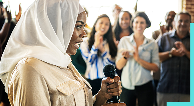 Photo d’une personne en chemise à col beige portant un foulard sur la tête, qui tient un microphone et fait un discours à un public en train d’applaudir dans une salle très lumineuse