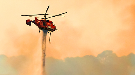 A photo of a red helicopter dropping fire retardant onto a blaze in a smoky orange-tinted sky