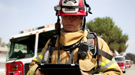 A photo of a first responder in a red helmet standing outside in front of a row of fire engines and using a tablet
