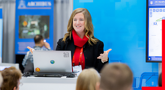 A presenter wearing a black coat and red scarf speaking to a crowd in the foreground in front of a blue screen at a conference exhibit
