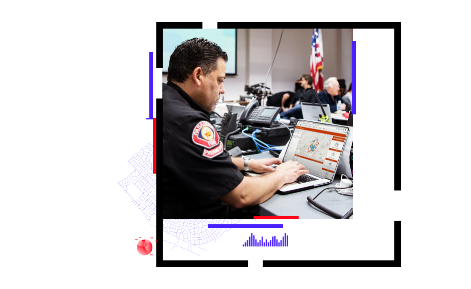 A photo of a police officer working on a laptop in a crowded conference room with other officers in the background