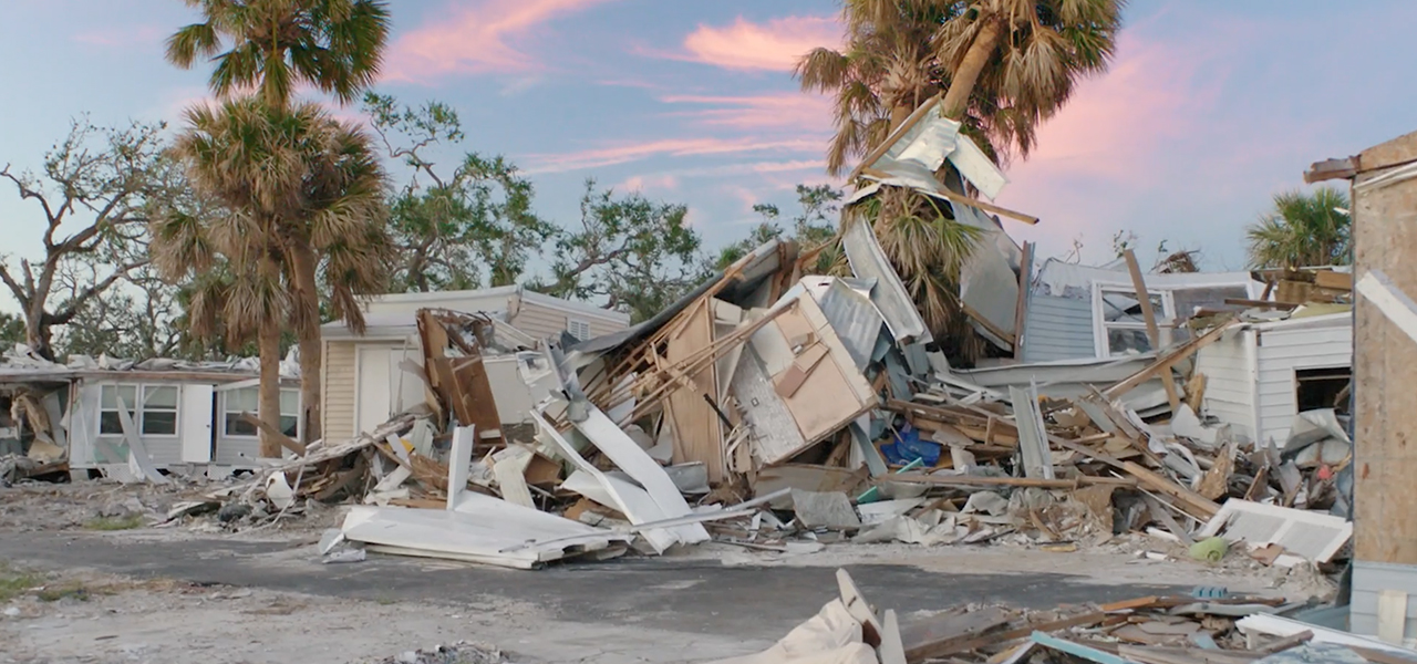 Mangled manufactured homes and other debris near palm trees under a blue sky