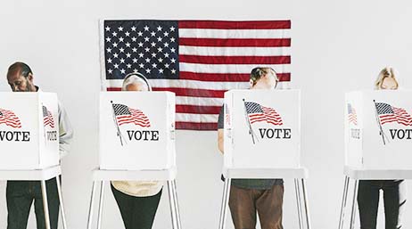 A photo of a row of voters standing within white voting booths, each printed with a US flag in red and blue and the word “VOTE”, with a US flag hanging on a white wall in the background