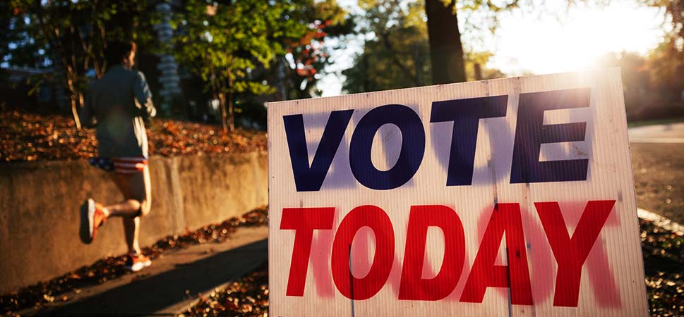 A photo of a white sign with the words “VOTE TODAY” in blue and red letters sitting on a tree-lined sidewalk as a person in shorts and tee shirt jogs past with a bright sunrise in the background