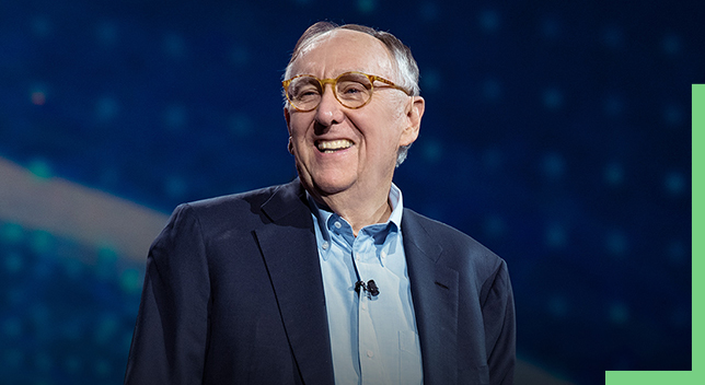 Una foto del fundador de Esri, Jack Dangermond, con camisa azul y chaqueta, sonriendo en el escenario de una Conferencia de usuarios de Esri