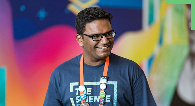 Uma pessoa sorridente em uma camiseta azul e um cordão de conferência em frente a um fundo abstrato colorido sem foco