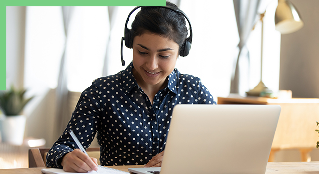 Foto einer lächelnden Person in einer blau-weiß gepunkteten Bluse, die ein Headset trägt und in einem sonnenbeschienenen Büro an einem Tisch sitzt und etwas auf einen Notizblock schreibt