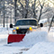 A white truck with a red plow attachment driving through a deep white snowdrift on a narrow tree-lined road through a park