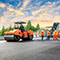A crew wearing orange safety vests work alongside heavy machinery to pave a new highway beneath a gold and blue sunrise