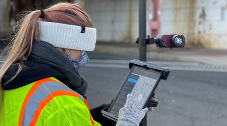 A mobile worker wearing knit gloves, a face covering, and a neon yellow vest working on a tablet
