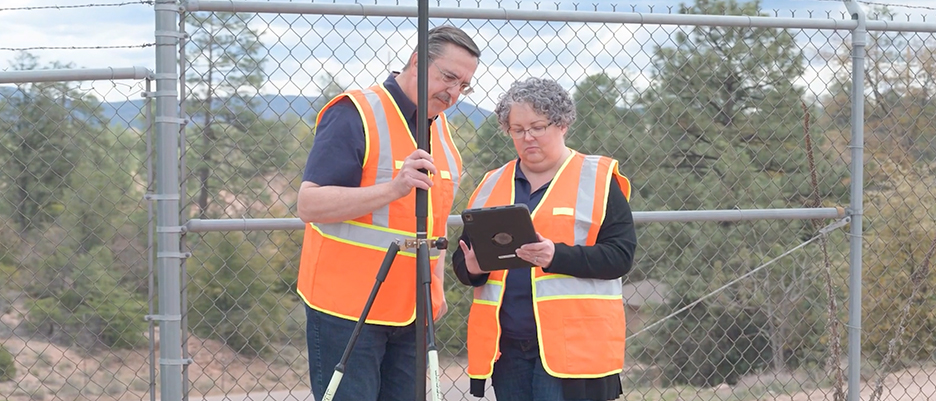 Two people wearing orange safety vests standing outside beside a chain-link fence sharing a tablet. A play button overlaid on the image indicates that the video can be played by clicking on the image