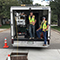 Two workers wearing yellow visibility safety vests standing among tools in the back of a box truck on a residential street