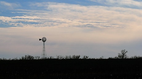 A photo of a large flat plain with a single windmill silhouetted against an overcast pale blue evening sky