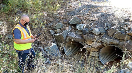 A photo of a person wearing a mask and yellow utility vest, holding a tablet, standing next to a large drainage area covered in rocks