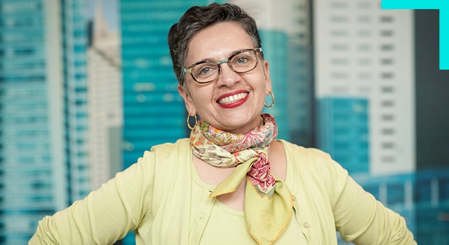 Une femme souriante portant un pull jaune et des lunettes pose avec des gratte-ciel en arrière-plan